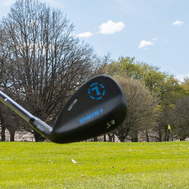 A Lazrus 60-degree wedge held just above short grass on a course, trees and a flag in the background, for chipping drills you can do at home this winter